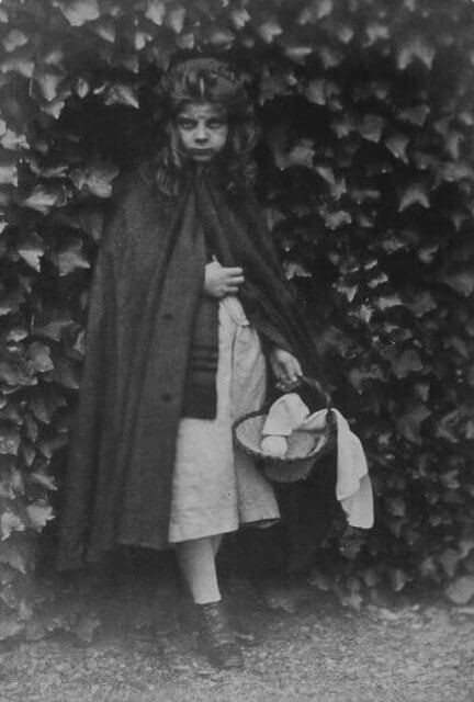 Photograph of Agnes Grace Weld as Little Red Riding Hood. With her right hand she holds together her cape, in the left hand she carries a basket.